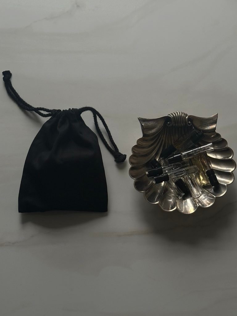 Scalloped silver shell dish holding miniature perfume bottles beside a black pouch on a pale stone surface.