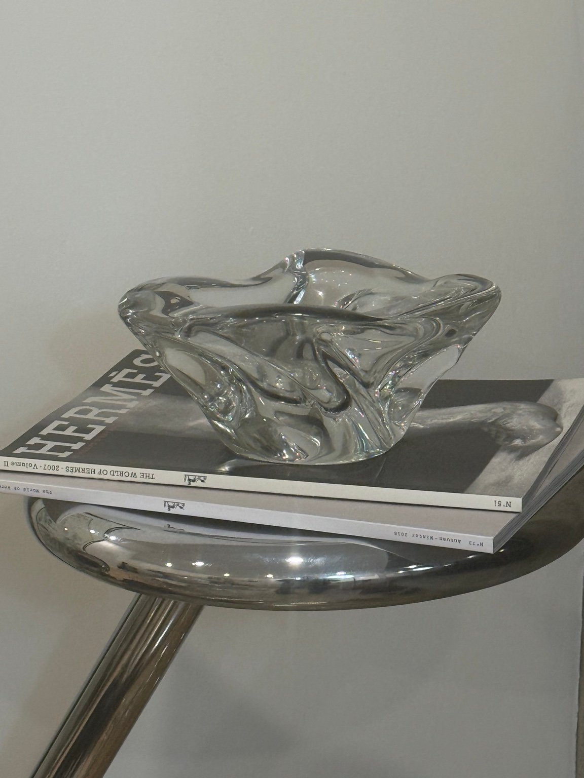 Sculptural clear glass ashtray on a book atop a polished metal side table.